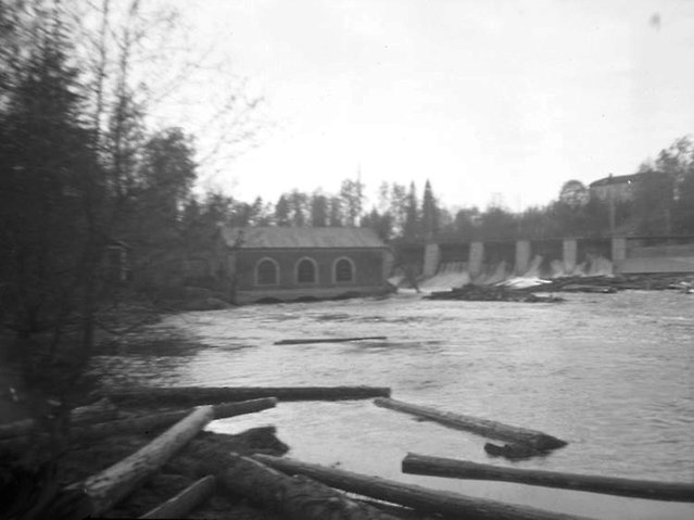 Svartvit bild. timmer i vatten framför kraftstation.
